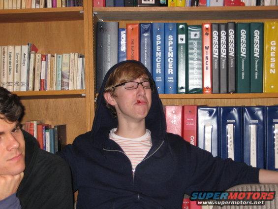 Two young men sit in front of bookshelves, one with a hood up