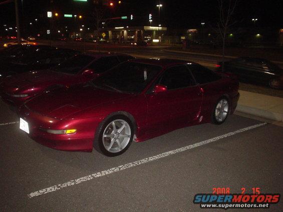 Red coupe parked in a lot at night, another red coupe behind it, and a dark car in the background