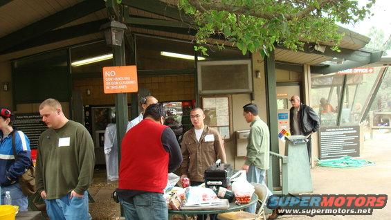 People stand around a table with a portable grill and food items outside a building with a "NO GUN HANDLING" sign