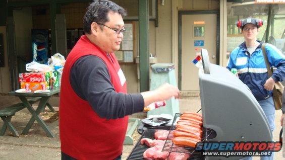 Man in red vest grilling hot dogs and meat on a barbecue with a picnic table and drinks in the background