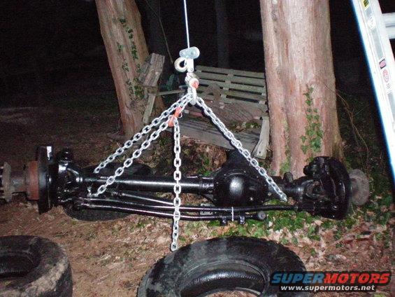 Black painted front axle assembly with chains attached, resting on the ground near trees and tires