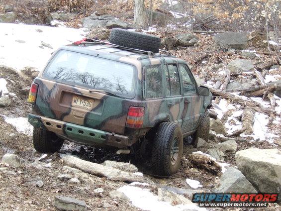 Camouflage Jeep Grand Cherokee with spare tire on roof, off-roading on rocky terrain