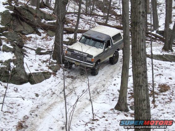 1987 Ford Bronco driving on a snowy trail between trees. Text on windshield reads 'THINK OF LOW PLACES'
