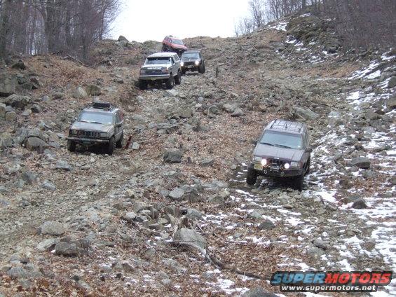 Several SUVs driving up a steep, rocky, wooded slope with some snow on the ground