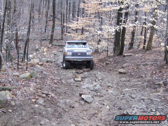 White pickup truck with lifted suspension driving on a rocky, muddy trail in a forest