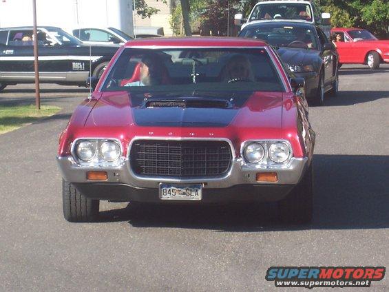 Red 1972 Ford Gran Torino Sport with black hood scoop, followed by a black Mustang and a red car