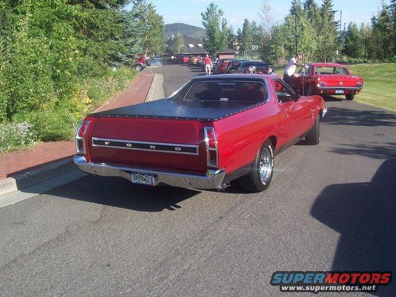 Red Ford Ranchero pickup truck with black tonneau cover, seen from rear. Another red car, possibly a Mustang, is behind it