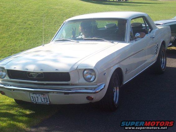 White 1965 Ford Mustang coupe parked on asphalt with grass in background. Driver's hand visible making peace sign