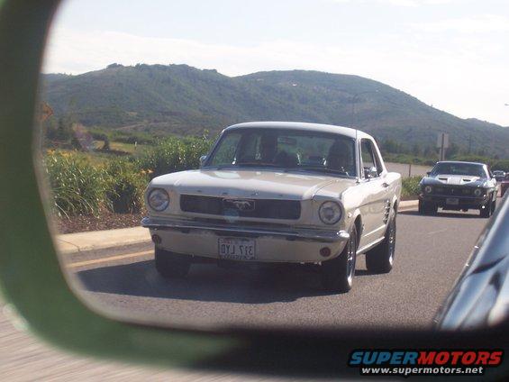 Cream-colored 1965 Ford Mustang driving on road, seen in side mirror. Another Mustang is behind it