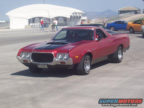 Red Ford Ranchero with black hood stripe parked outside. Other cars and people in background