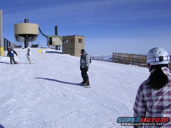 Snowboarders on a ski slope with a ski lift and buildings under a clear blue sky