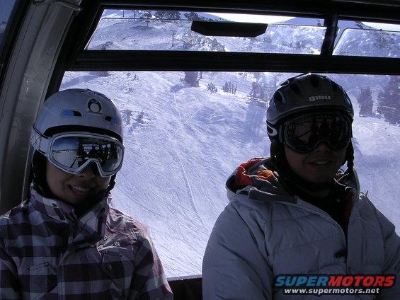 Two people in ski gear, wearing helmets and goggles, ride in a ski lift with a snowy mountain slope visible outside