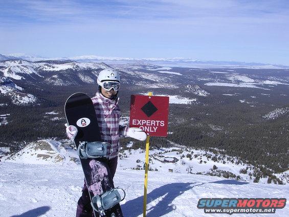 Snowboarder in purple plaid jacket and goggles holds snowboard near a red diamond sign that reads EXPERTS ONLY