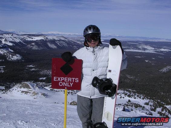 Snowboarder in white jacket and helmet holding a snowboard, standing next to an 'EXPERTS ONLY' sign on a snowy mountain