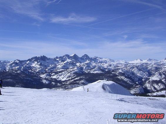 Snowy mountains under a blue sky with ski slopes and a few skiers visible. Watermark says SUPERMOTORS