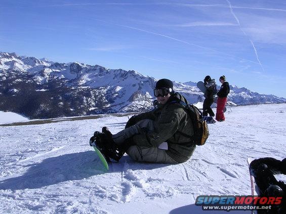Snowboarder sitting in snow with mountains behind. Supermotors.net logo visible