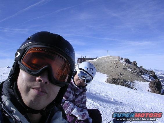 Two skiers wearing helmets and goggles on a snowy mountain with a building in the background
