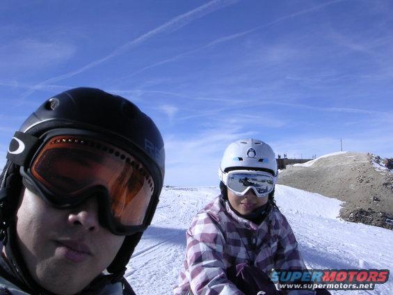 Two people in ski gear on a snowy slope, one in foreground wearing black helmet and orange goggles, the other in background wearing white helmet and goggles