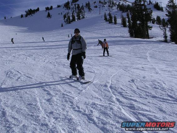 Person snowboarding down a snowy mountain slope with trees and ski lifts in the background