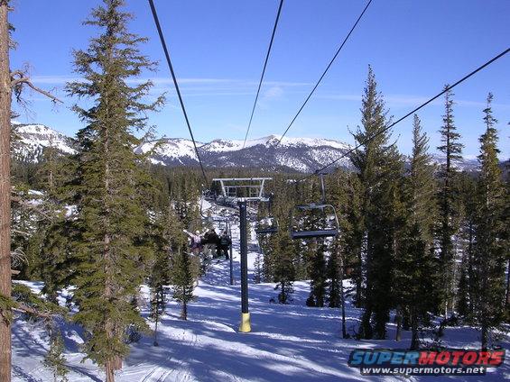Ski lift chairs ascend a snowy mountain slope with pine trees and snow-capped peaks under a clear blue sky