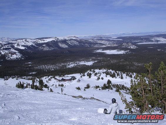 Snowboarder in white jacket sits on snowy slope overlooking a vast mountain range and forest