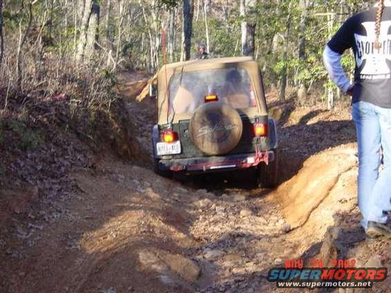 Tan soft-top Jeep Wrangler on a dirt trail, rear view