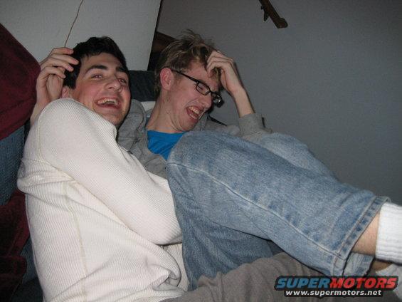 Two young men laughing, one in a white waffle-knit shirt, the other in a blue shirt and jeans. Supermotors.net logo in lower right