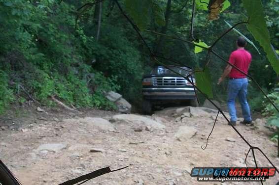 1995 Ford Bronco driving on a dirt and rock path in a wooded area