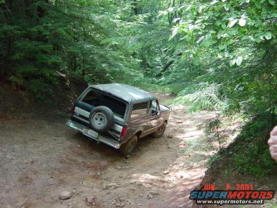 Brown and silver Ford Bronco on a dirt trail, rear view, spare tire mounted on back