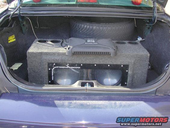 Dark blue 1999 Ford Crown Victoria trunk interior with a large subwoofer box, two subwoofers, an amplifier, and a spare tire