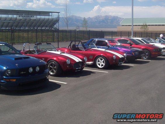 Several sports cars, including a blue Mustang and red Cobras, parked in a lot with a grandstand in the background