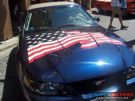 Blue convertible with American flag decal on hood, parked on street. Another red convertible visible behind it