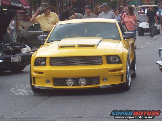 Yellow Ford Mustang with a hood scoop and aftermarket grille, seen from the front