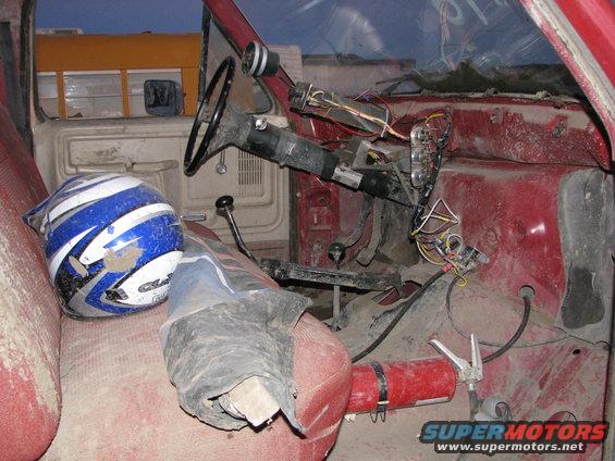 Interior of a red truck, showing the dashboard, steering wheel, and exposed wiring. A blue and white helmet is on the seat