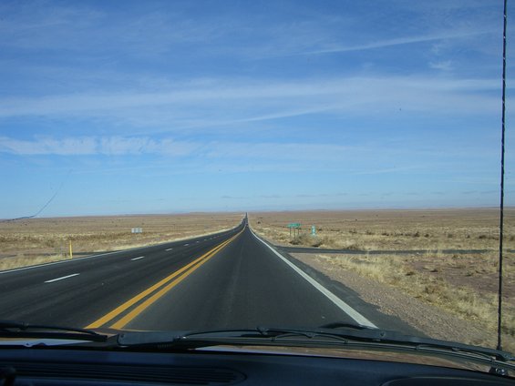 Desert highway stretching to the horizon under a blue sky with scattered clouds. Dry grass lines the roadside