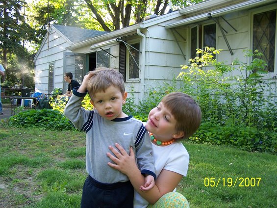 Young boy with hand on head, being held by a girl, with a house and trees in the background