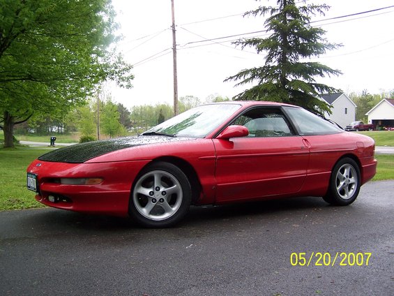 Red 1996 Ford Probe coupe with a black hood, parked on a wet asphalt driveway