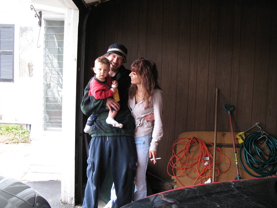 Man holding a baby, woman standing next to him, in a garage with tools and supplies