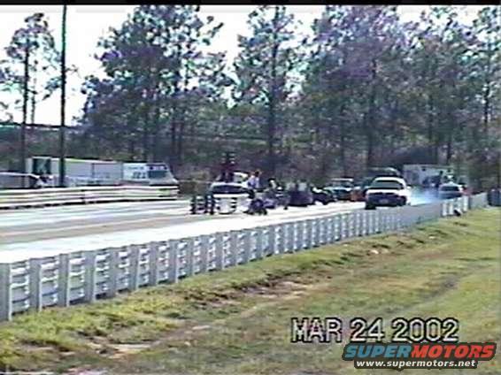 Drag strip with cars lined up and one accelerating, trees and trailers in background. Watermark reads MAR 24 2002 SUPERMOTORS