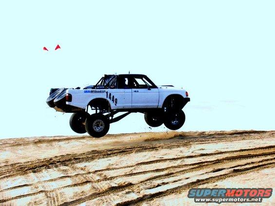White pickup truck with roll cage and large tires airborne over sand dunes. Number 1449 visible on side