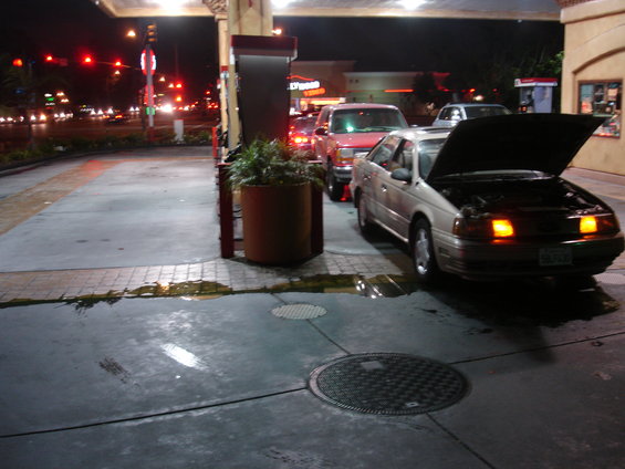 Beige 1991 Ford Taurus with hood open and hazard lights on at a gas station