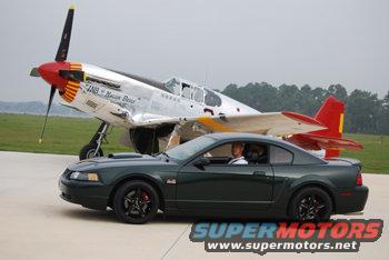 Dark green 2001 Ford Mustang parked next to a P-51 Mustang airplane on tarmac