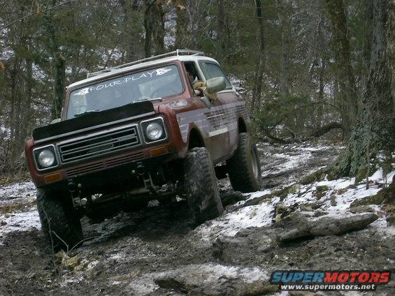Red International Harvester Scout II off-roading on a muddy, snowy trail in the woods. Windshield sticker says "FOUR PLAY"