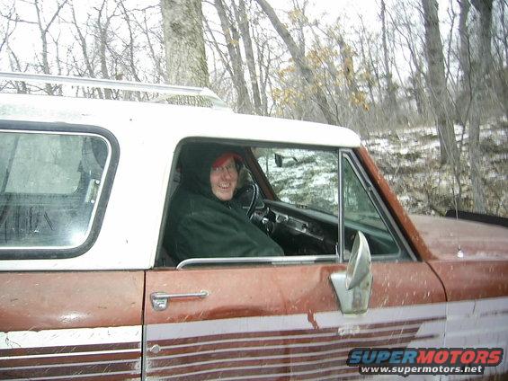 Person wearing a red hat and green jacket inside a rusty SUV with a white roof and rack. Trees and snow in background