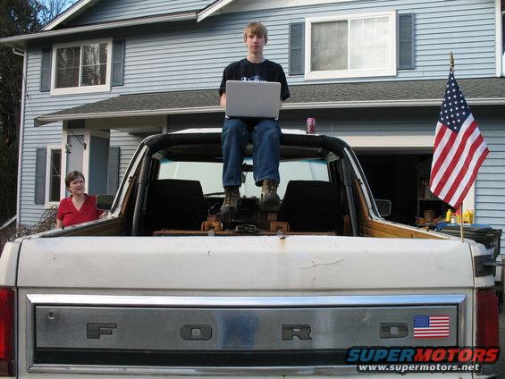 White Ford pickup truck rear view, person sitting on tailgate with laptop, American flag on fender