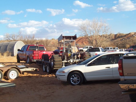 Red pickup truck towing a large off-road vehicle with paddle tires. White sedan and other trucks are parked nearby