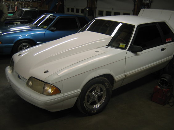 White 1992 Ford Mustang coupe with a large aftermarket hood, drag racing tires, and a red decal on the door