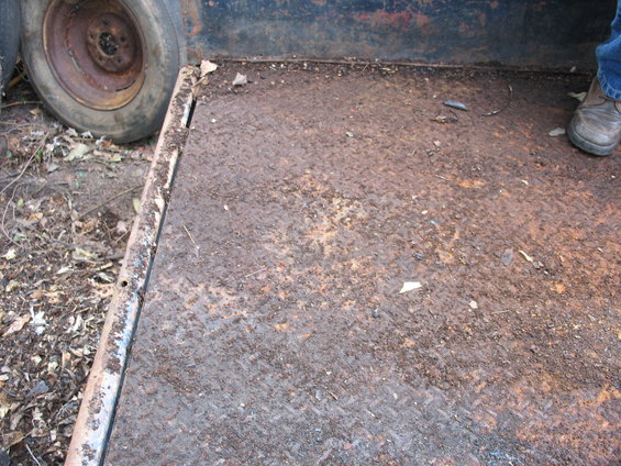 Rusted diamond plate trailer bed with a spare tire visible on the left. A person's shoe is visible on the right