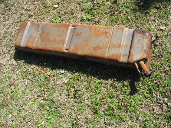 Rusty metal fuel tank lying on grass. It has several raised horizontal bands and two pipe fittings on one end