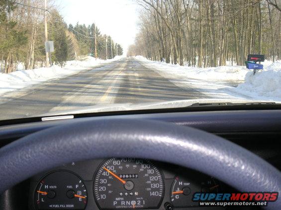 View from inside a car, looking out at a snowy road ahead. Steering wheel and dashboard visible